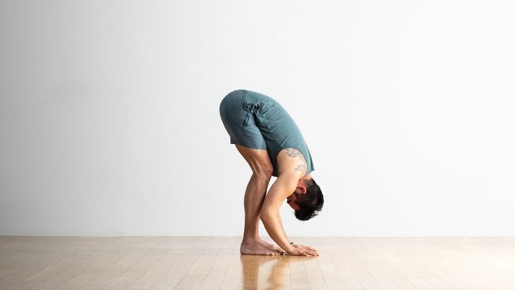 Un hombre con cabello oscuro se inclina hacia adelante en Uttanasana, de pie se pliega hacia adelante. Lleva pantalones cortos y top azul grisáceo. Sus rodillas están ligeramente dobladas. Él tiene sus manos en el piso de madera cerca de sus pies.